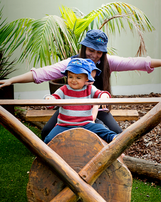 Column image Early childhood centre educator helping toddler balance on outdoor wooden play equipment